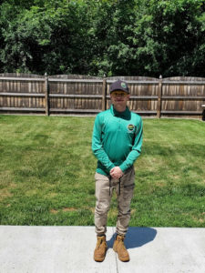 A technician dressed in a green long-sleeved shirt and khaki pants poses for a picture in a New York backyard.